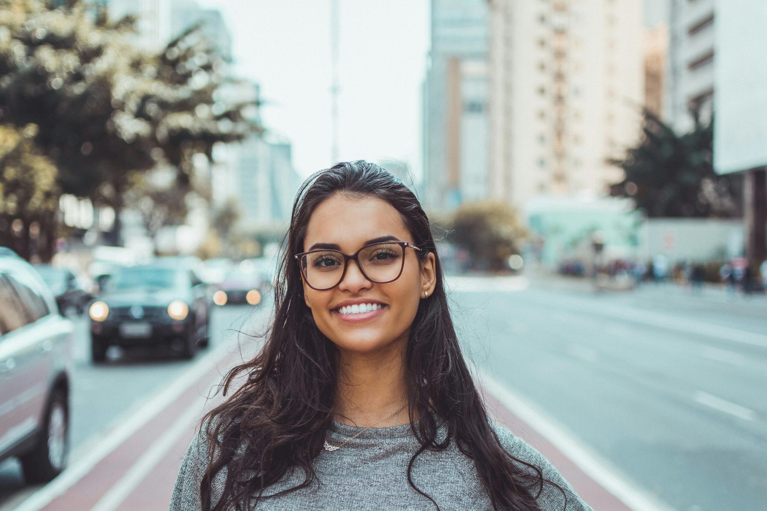 A young woman with glasses smiling on a city street, embracing urban lifestyle.